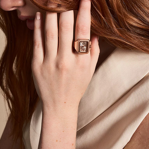Rose gold-tone watch ring with rectangular face, Roman numerals, adjustable band on hand